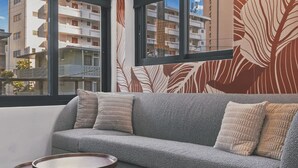 A modern living room with a grey sofa, two round wooden side tables, and a large window with a view of buildings. - The Walk-Up Waikiki Beach  (Honolulu)
