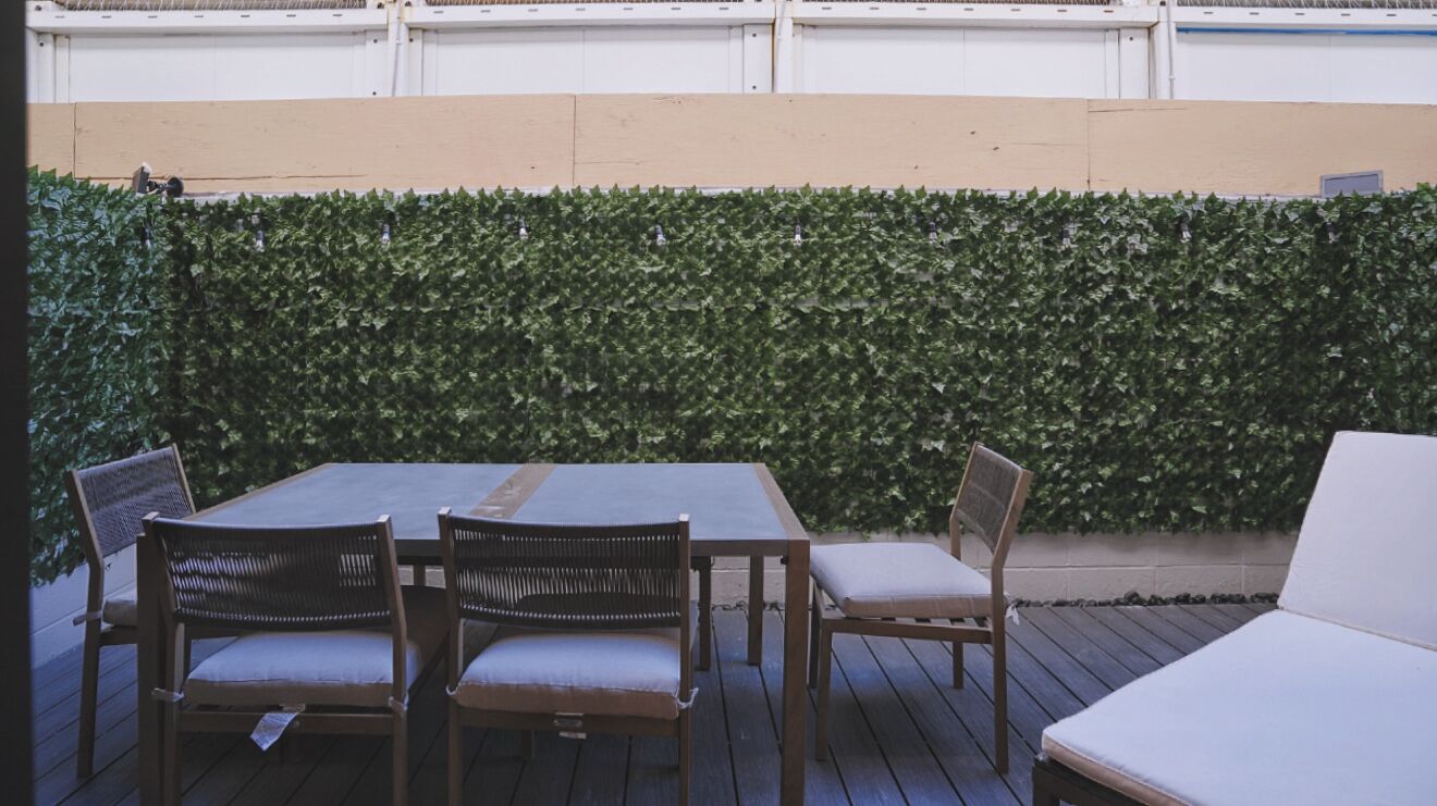 Eine Terrasse im Freien mit einem Holztisch und -stühlen, einer weißen gepolsterten Bank und einer grünen Heckenwand.