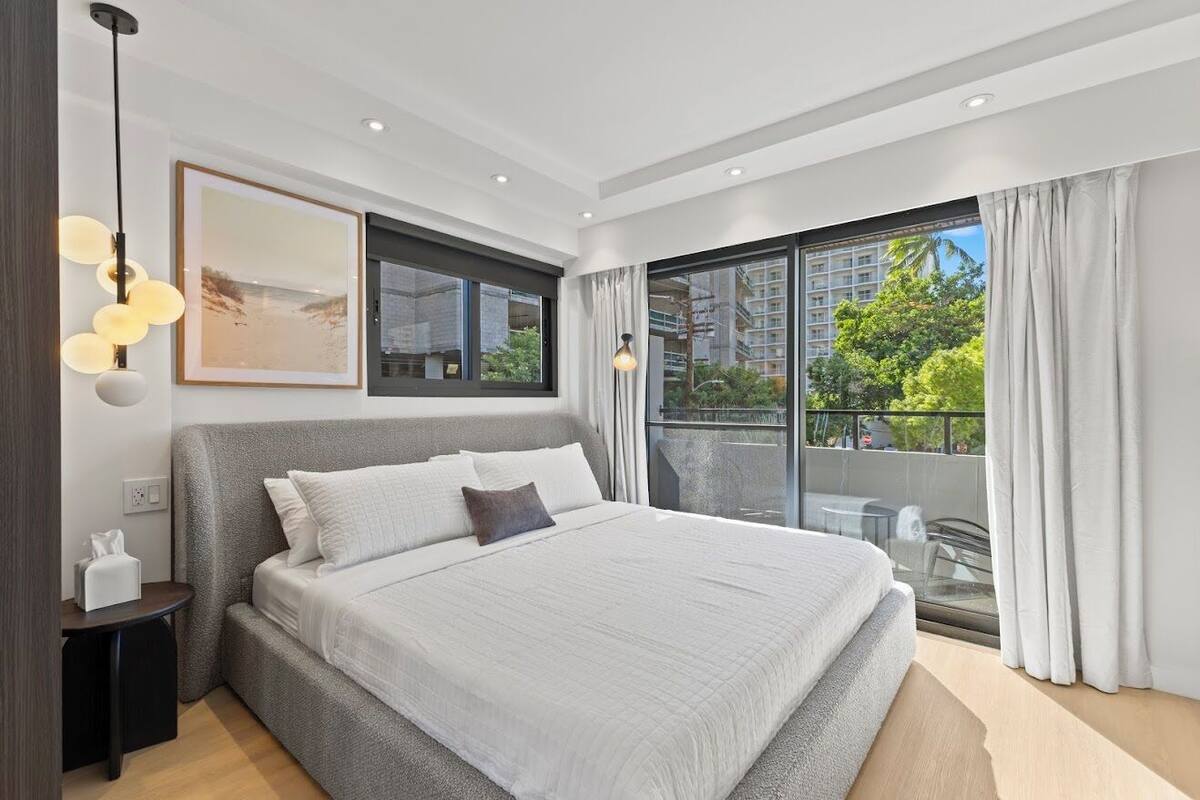 Une chambre moderne avec un grand lit, une vue sur la verdure et les bâtiments depuis un balcon, et un tableau encadré au mur.