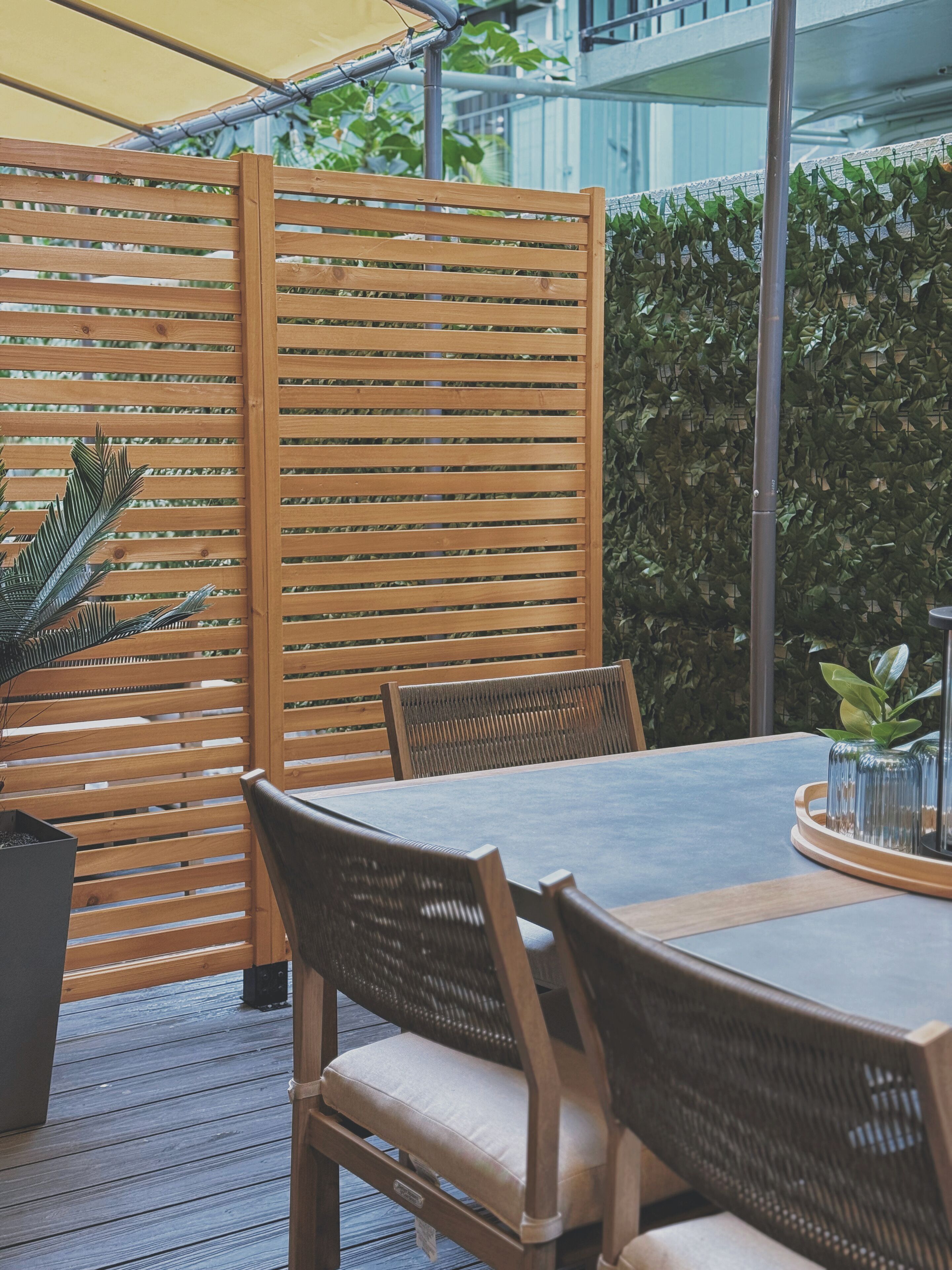 A wooden outdoor seating area with a table and chairs, surrounded by a bamboo fence and greenery.