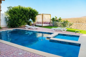 Une piscine divisée en deux zones, entourée d’une clôture, avec un espace de repos couvert et des chaises longues.