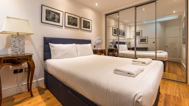 A modern bedroom with a large bed, a bedside table with a lamp, and a mirror. - Spacious 4 Bed Apartment Opposite British Museum (London)
