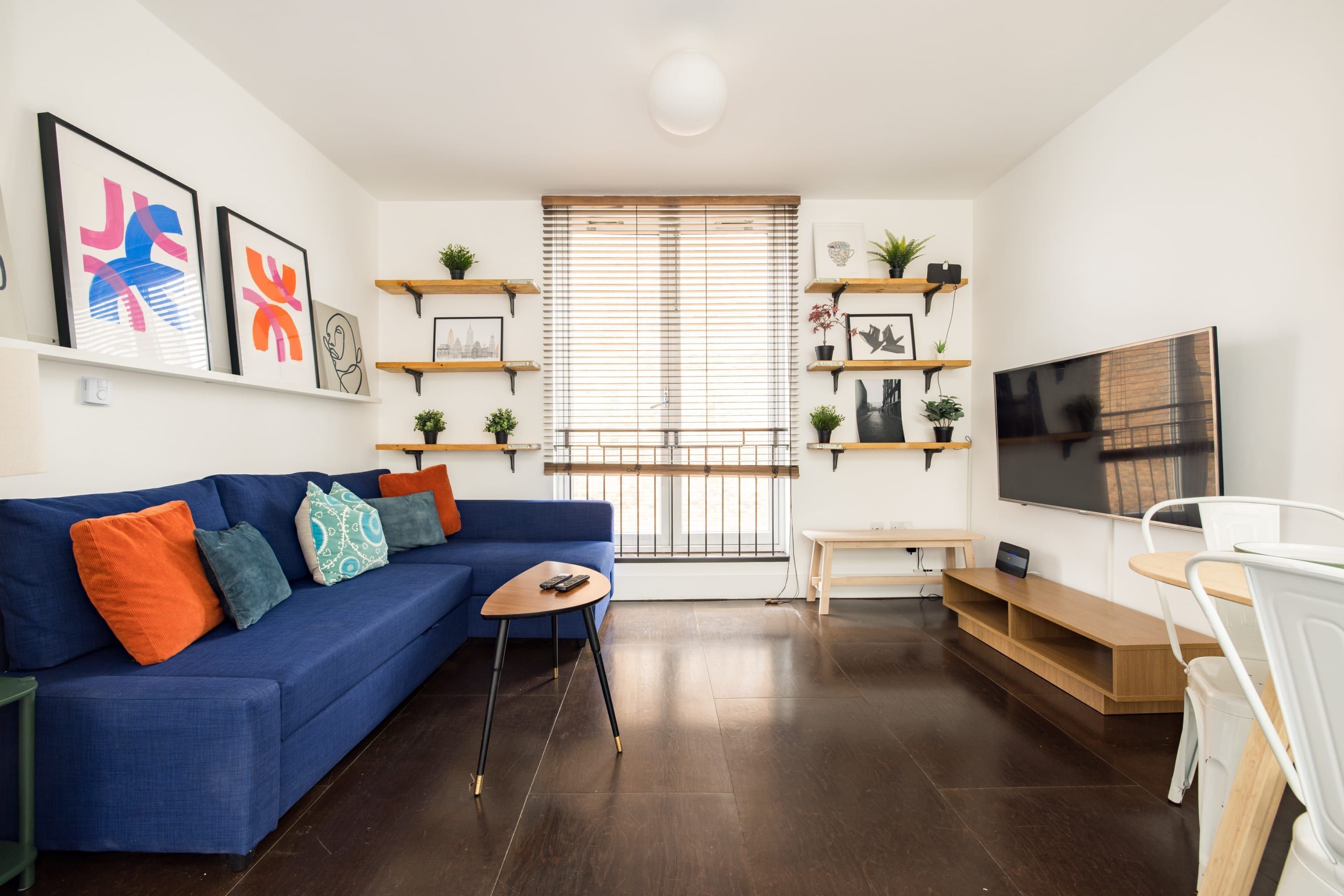 A modern living room with a blue sofa, wooden shelves with plants, a TV, and a dining area.