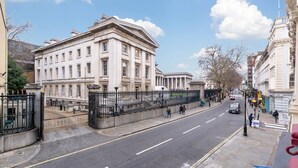 A street view with a large classical building, a clear sky, and a few pedestrians.