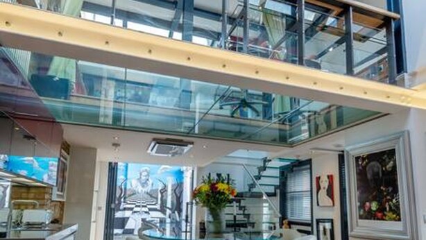 A modern interior with a glass staircase, a dining area with a round table and bar stools, and a kitchen visible in the background.