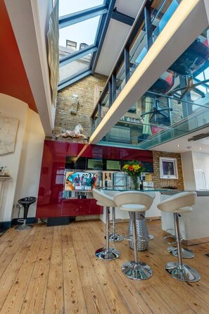 A modern interior with a wooden floor, a bar area with stools, and a view of the city through large windows.