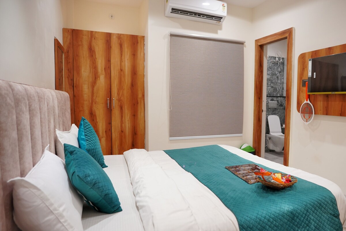Un dormitorio con cama, armario, ventana con persianas, zona de ducha y televisor montado.
