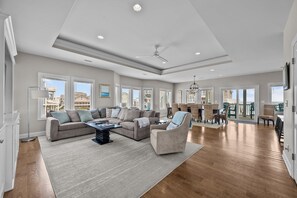 A spacious living room with a large sectional sofa, a coffee table, and a fireplace. There are multiple windows providing natural light and a view of the outside.