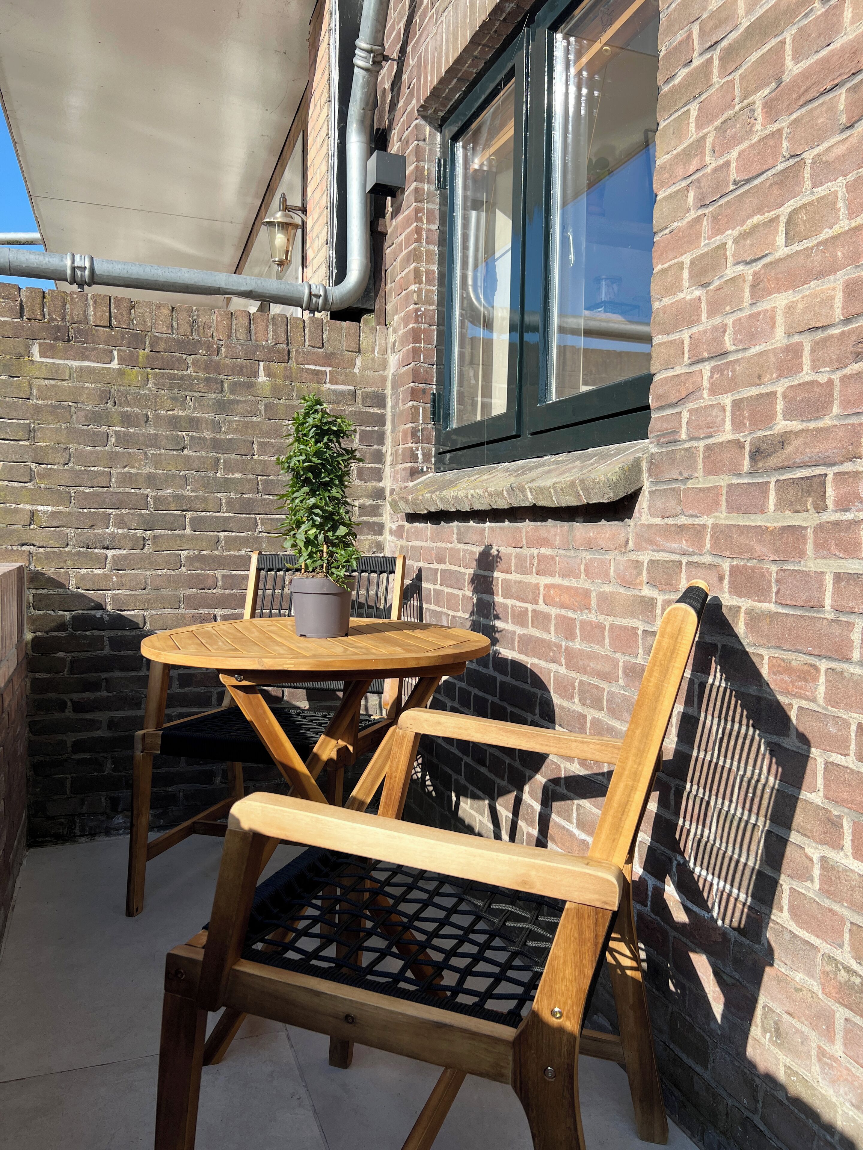Een houten tafel met stoelen geplaatst tegen een bakstenen muur, met een raam erboven en een bloempot naast de tafel.