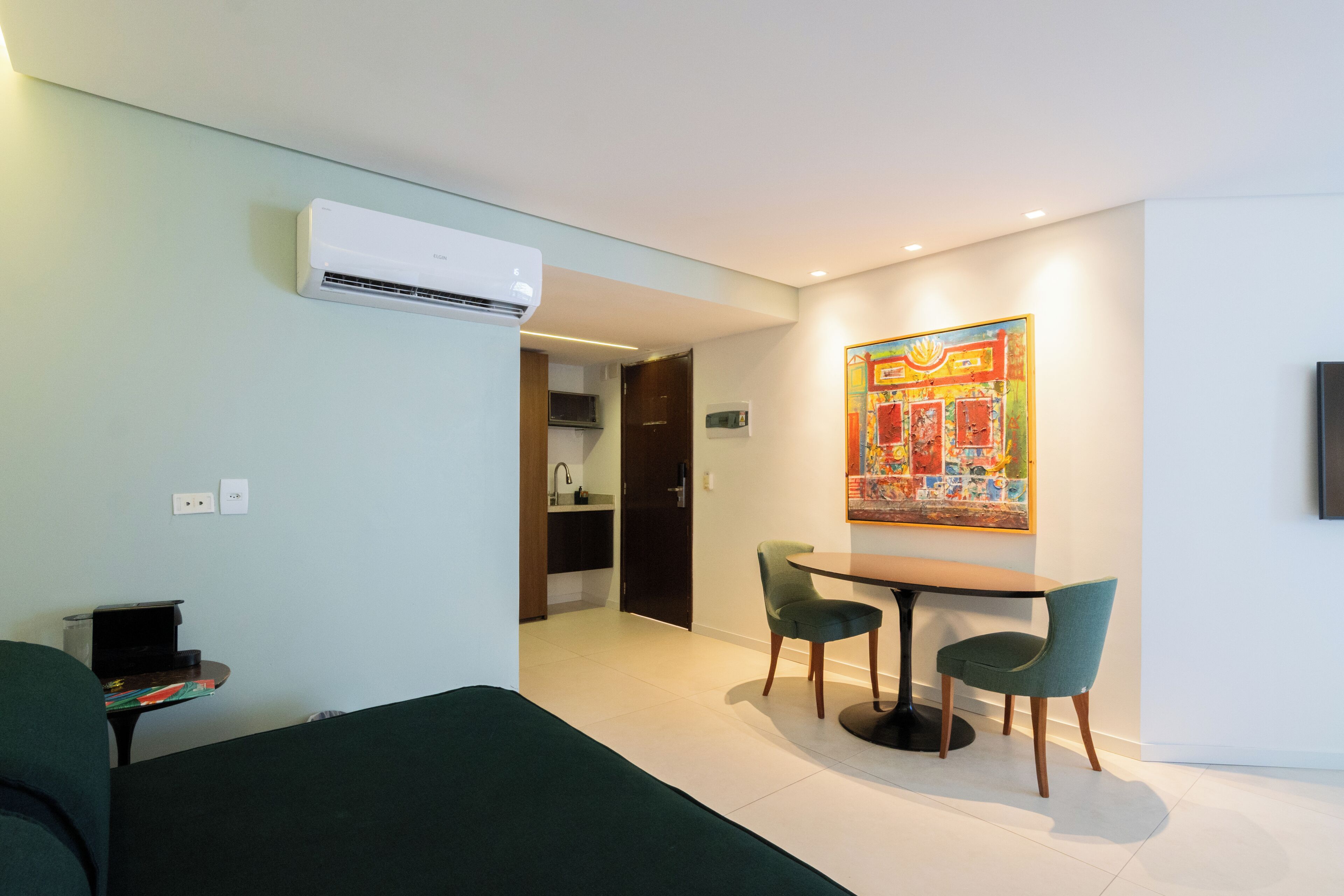 A hotel room with a bed, a dining table with chairs, an air conditioning unit, a painting on the wall, and a kitchen area visible through an open door.