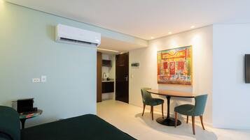 A hotel room with a bed, a dining table with chairs, an air conditioning unit, a painting on the wall, and a kitchen area visible through an open door.