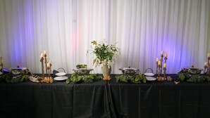 Una mesa de buffet con platos cubiertos, velas y follaje, sobre un fondo de cortinas blancas.