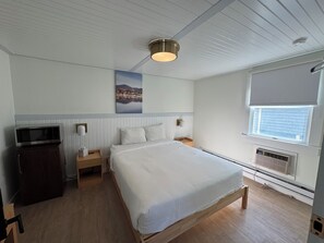 A bedroom with a bed, a nightstand, a dresser, a wall-mounted lamp, a ceiling light, a window with blinds, and a radiator. - Queen Room - SQR (Lake Placid)