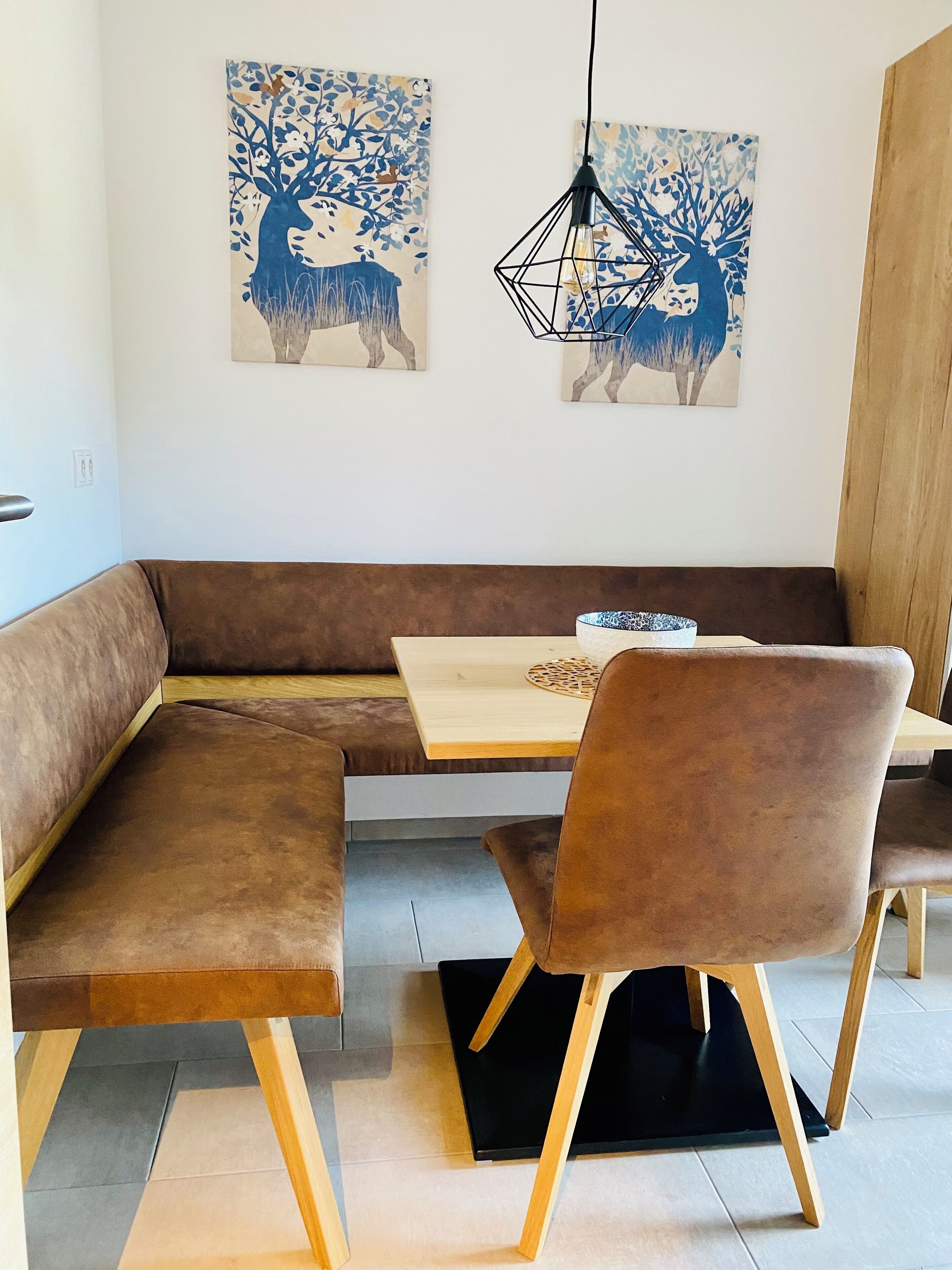 Un espace repas moderne avec une table en bois, des bancs rembourrés de couleur marron et deux tableaux représentant des cerfs, accrochés au mur.