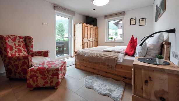A bedroom with a bed, a wooden wardrobe, a chair, and a desk with a plant.