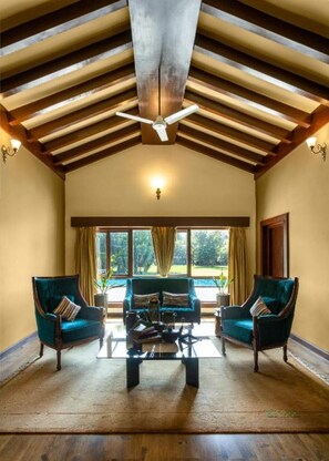 Een ruime kamer met een hoge plafond, een plafondventilator, een groot raam met gordijnen, en twee fauteuils met een salontafel in het midden.