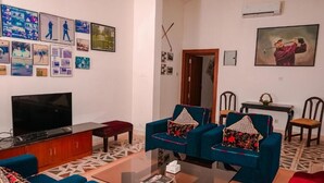 A living room with a flat-screen TV, two blue sofas with patterned cushions, a coffee table, and a wall decorated with various framed photos and a painting.