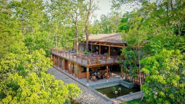 A wooden house with a balcony, surrounded by lush greenery and a small pond.