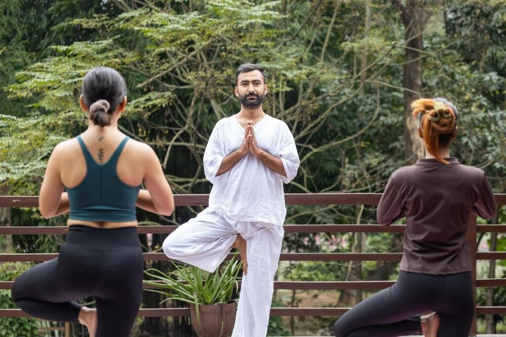 Three individuals practicing yoga outdoors with a wooden railing and greenery in the background.