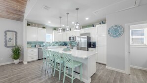 A modern kitchen with white cabinets, a large island with bar stools, pendant lighting, and a clock on the wall. - Spacious upscale home, community pool, private hot tub, close to the beach! (Port Aransas)
