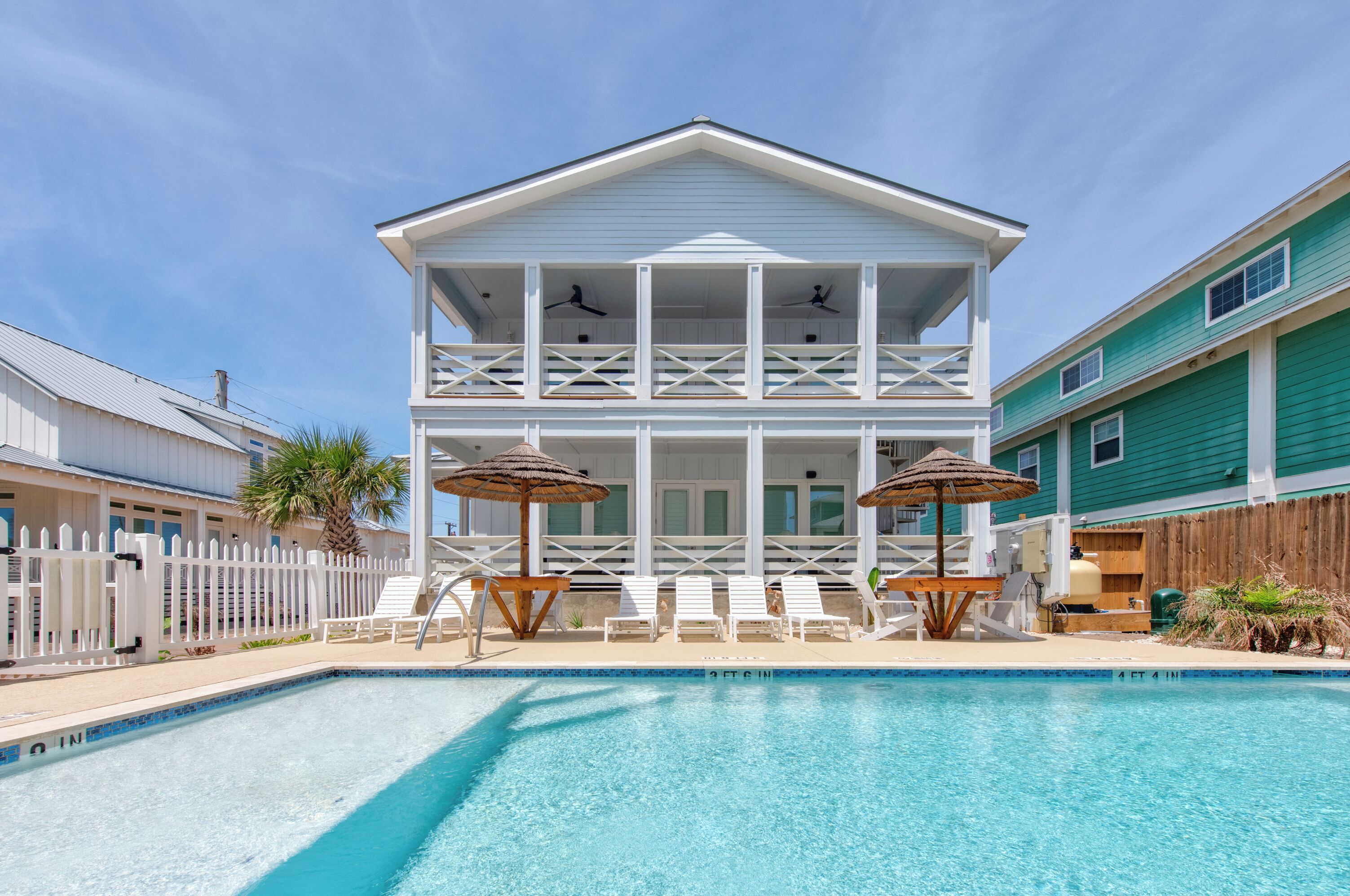 A two-story house with a pool, white railings, and a wooden deck with lounge chairs and umbrellas.