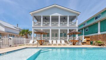 A two-story house with a pool, white railings, and a wooden deck with lounge chairs and umbrellas.