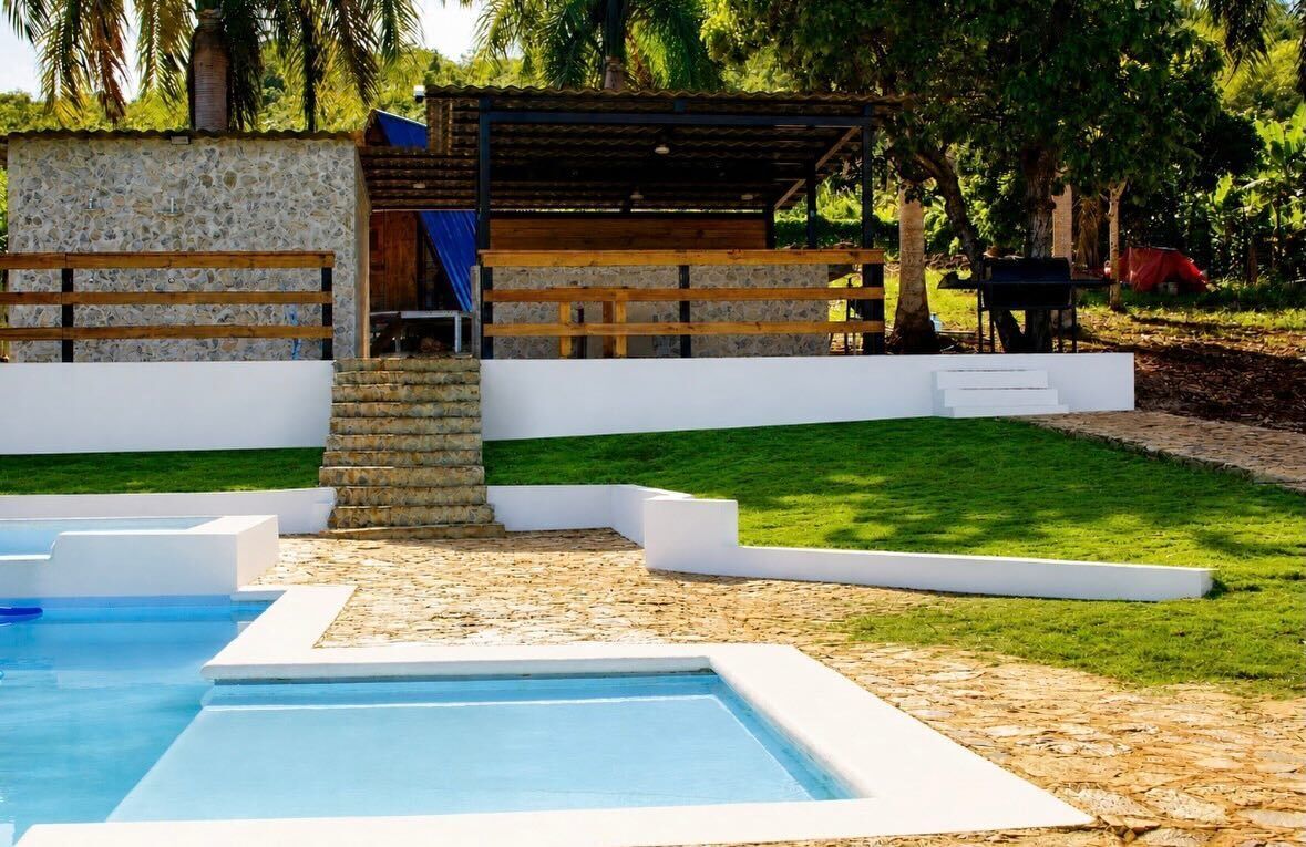 Une piscine bordée d’un mur en pierre et dotée d’une terrasse en bois, entourée d’une pelouse bien entretenue et d’arbres.