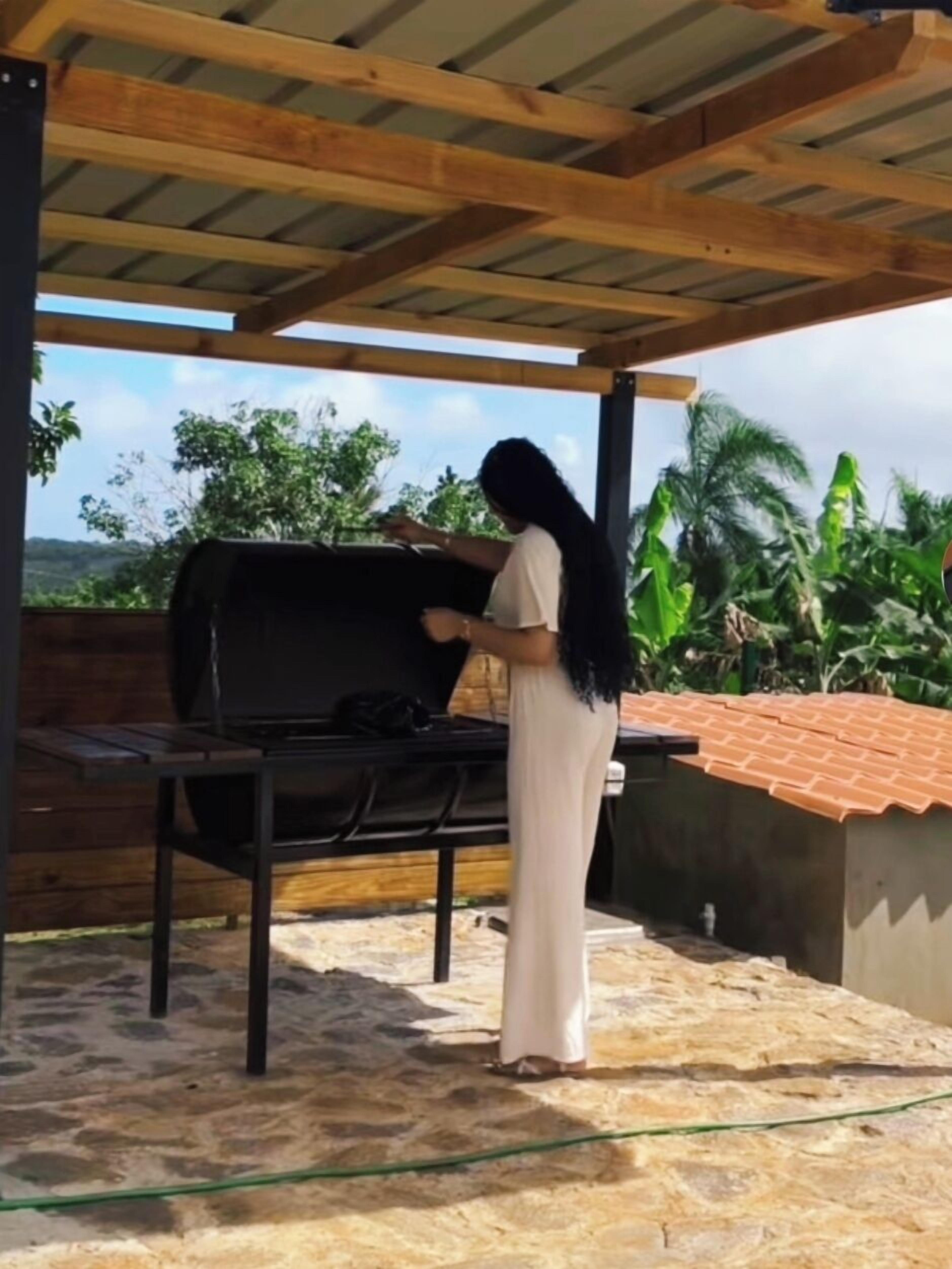 A covered outdoor area with a grill, a person standing nearby, and a view of greenery in the background.