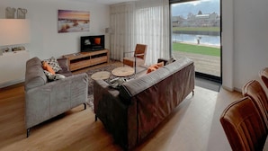 Un salon moderne avec un canapé gris, un canapé marron, une table basse en bois et un téléviseur posé sur un meuble en bois. Une grande fenêtre offre une vue sur un immeuble et un espace verdoyant.
