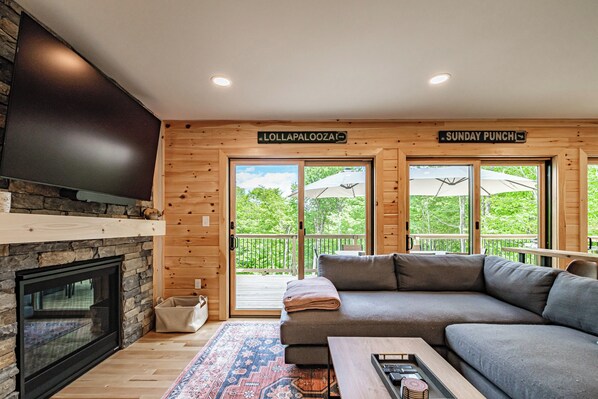A modern living room with a stone fireplace, a grey sofa, and a large TV. There are signs for 'Lollapalooza' and 'Sunday Punch' indicating nearby events.