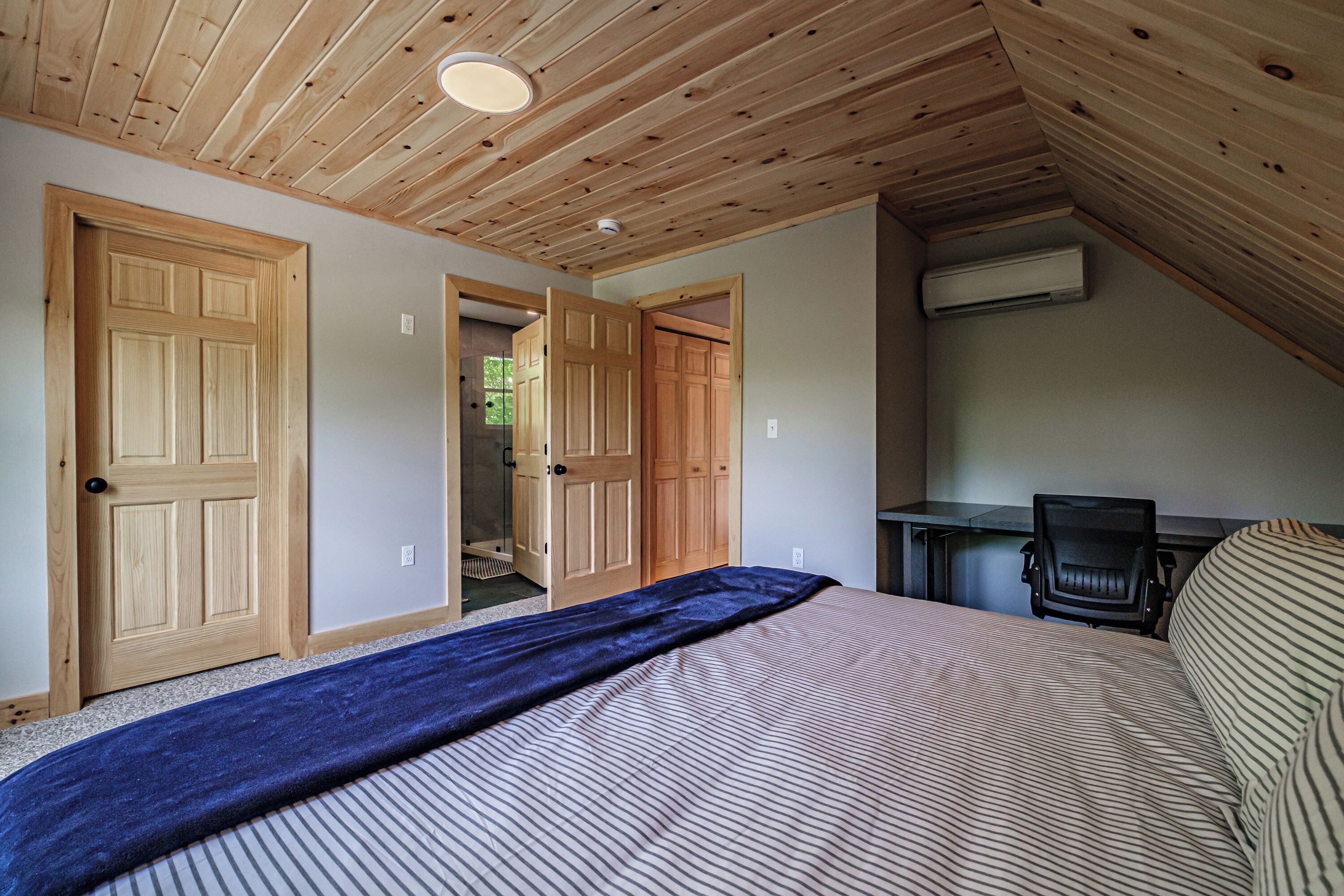 Ein Schlafzimmer mit schrägverdeckter Decke, Holztüren, einem Bett mit gestreifter Bettwäsche, einem Schreibtisch mit Stuhl und einer Klimaanlage.