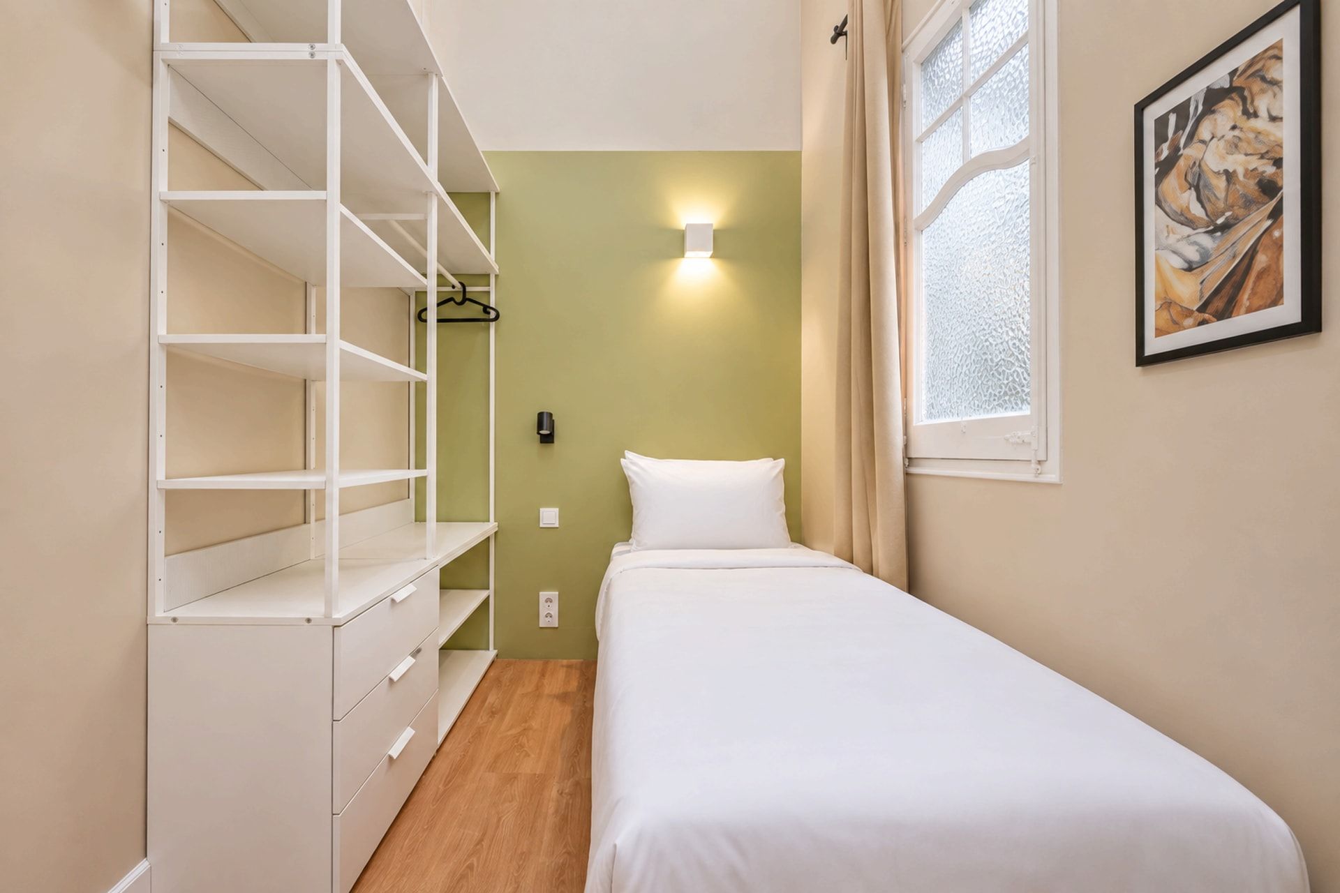 A small, neatly organized bedroom with a single bed, a wardrobe with multiple shelves, a wall-mounted light, and a window with curtains.