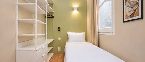 A small, neatly organized bedroom with a single bed, a wardrobe with multiple shelves, a wall-mounted light, and a window with curtains.