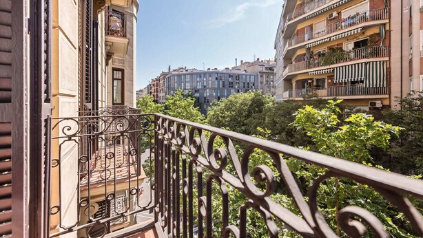 A balcony with wrought iron railings overlooking a cityscape with multi-story buildings and lush greenery. - Sweett | Sepulveda 2C (Barcelona)