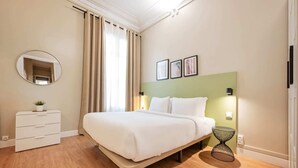 A hotel room with a bed, a bedside table, a mirror, a wall-mounted picture, and a window with curtains. - Sweett | Sepulveda 2C (Barcelona)