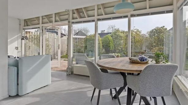 A modern dining area with a round table, grey upholstered chairs, and a view of the garden through large glass doors. - Villa Dune I (Ouddorp)