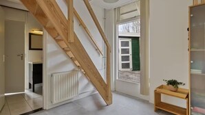 A staircase with wooden railings leading to an upper floor, a radiator on the wall, a window with a view of the outside, and a small wooden table with a plant. - Villa Dune I (Ouddorp)