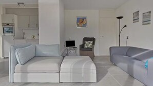 A modern living room with a grey sofa, a white sofa, and a grey armchair. There is a flat-screen TV on a small table, a painting on the wall, and a floor lamp. - Villa Dune I (Ouddorp)