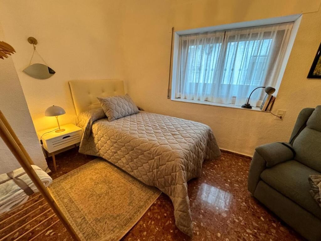 A bedroom with a bed, a nightstand, a lamp, a window with sheer curtains, a green armchair, and a small table with a lamp.