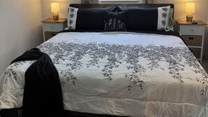 A neatly made bed with a floral bedspread, two bedside lamps, a ceiling fan, and a window with blinds.
