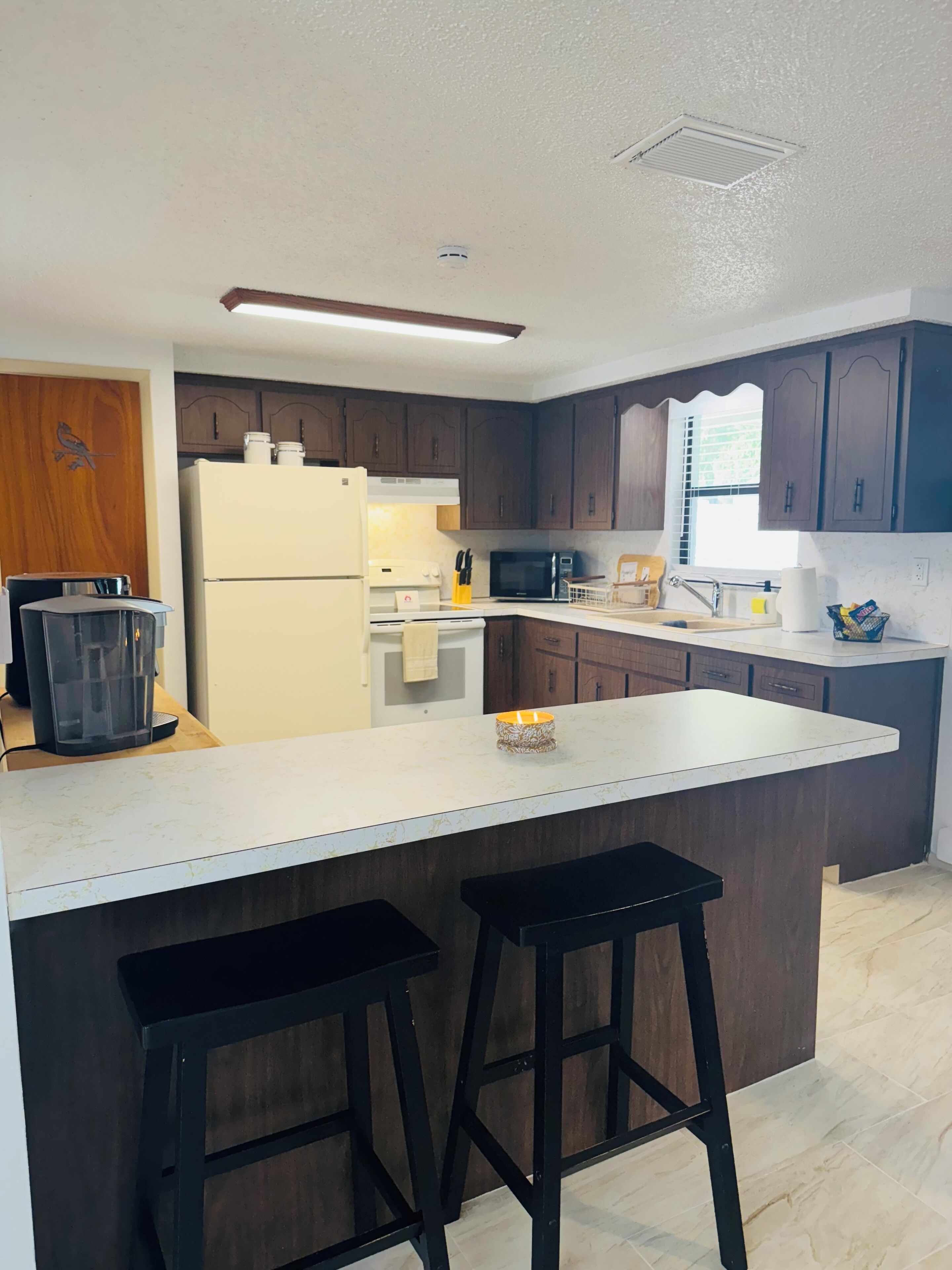 A modern kitchen with dark wooden cabinets, a white countertop, a built-in oven, a microwave, and a refrigerator. There are two black stools at the island.