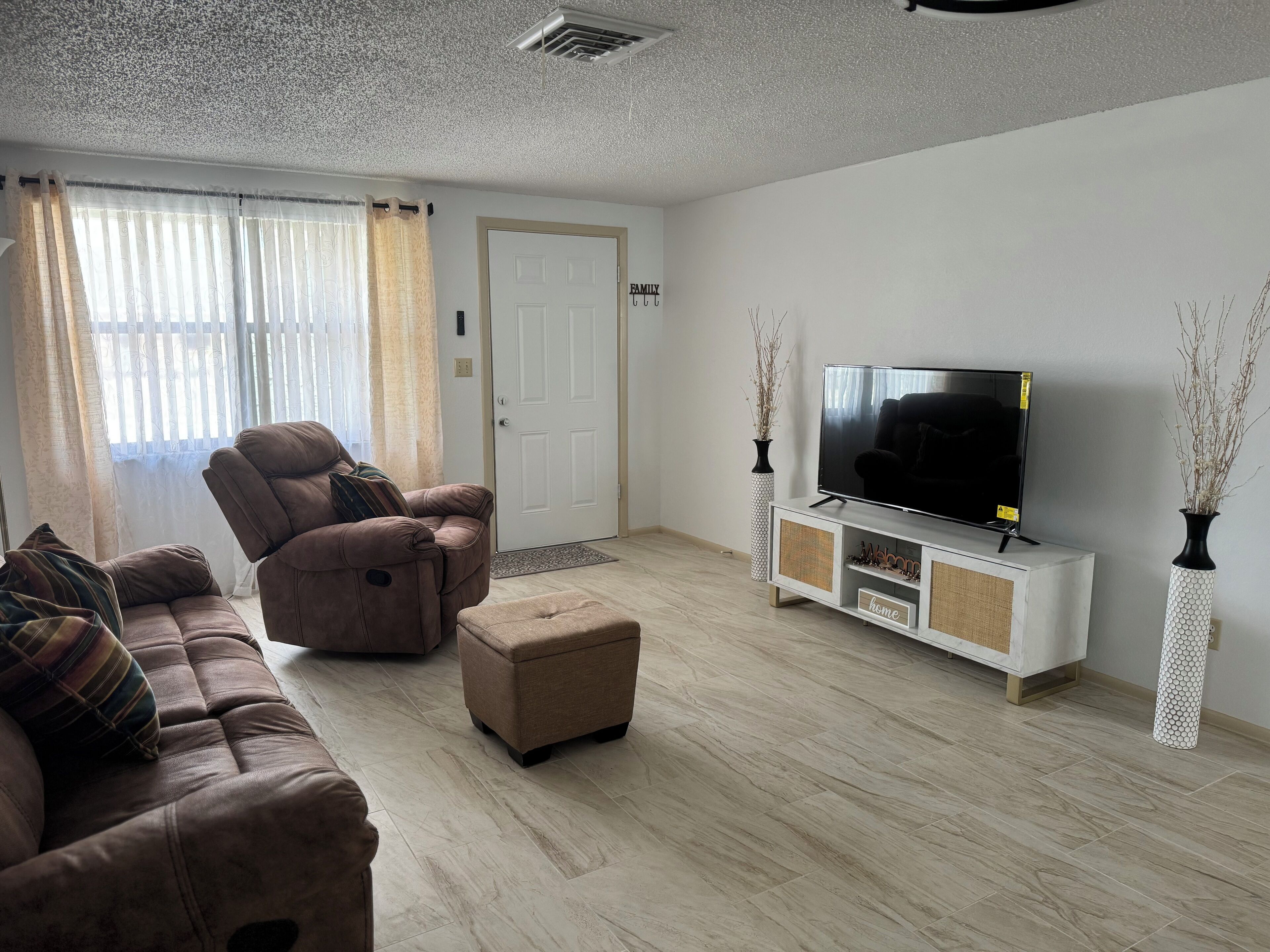 Una sala de estar con un sofá marrón, un sillón reclinable, un televisor sobre un mueble blanco y una puerta con un letrero que dice 'WELCOME'.