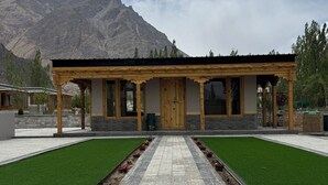 Un edificio con fachada de madera y detalles de piedra, un camino empedrado que lleva a la entrada, y montañas al fondo.