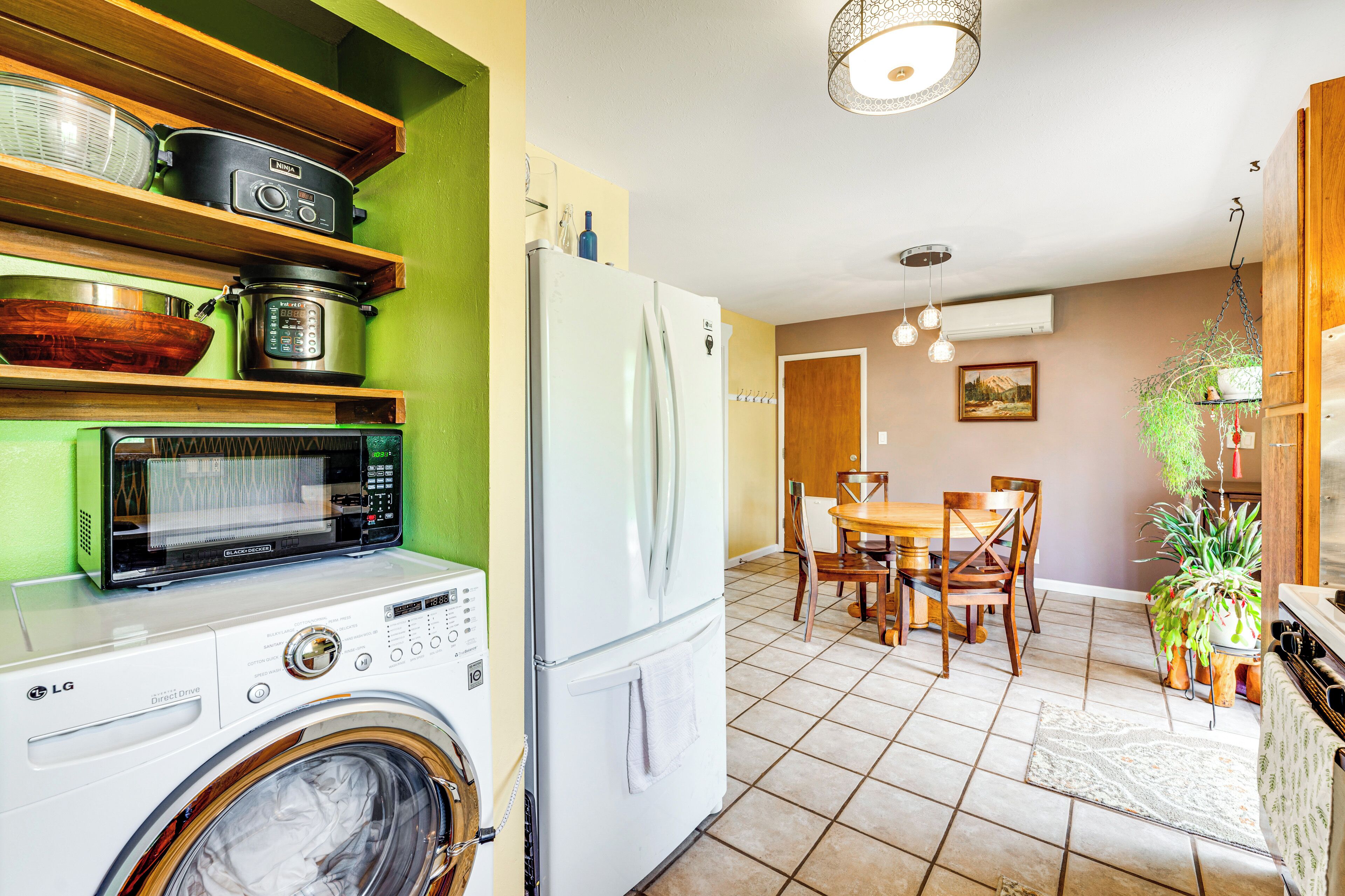 A compact kitchen with a washing machine, a refrigerator, and a microwave. There is a dining area with a table and chairs, and a hanging plant.