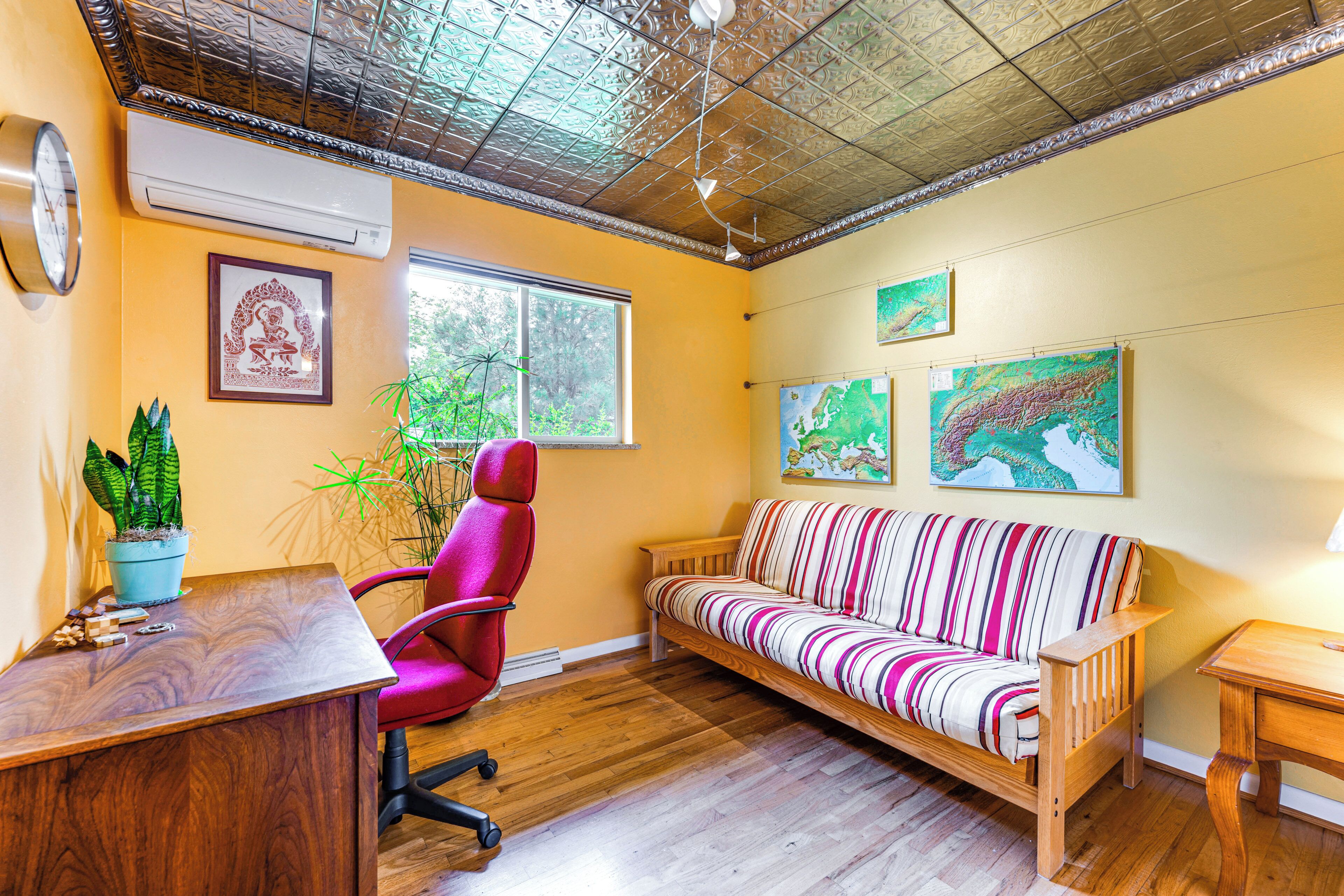 A room with a wooden desk, a red office chair, a striped sofa, and a small table. There is a clock on the wall and a potted plant on the desk.