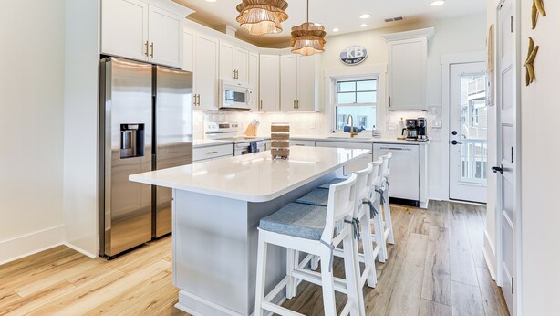 A modern kitchen with white cabinetry, a stainless steel refrigerator, a central island with bar stools, and pendant lighting. - Walk to the Waves: Coastal Kure Beach Retreat (Kure Beach)
