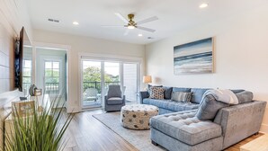 A spacious living room with a grey sectional sofa, a round ottoman, a flat-screen TV, and a ceiling fan. - Walk to the Waves: Coastal Kure Beach Retreat (Kure Beach)