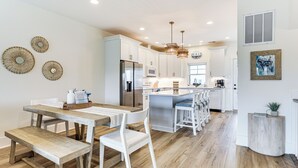 A modern kitchen with white cabinetry, a central island, and a dining area with a wooden table and benches. - Walk to the Waves: Coastal Kure Beach Retreat (Kure Beach)