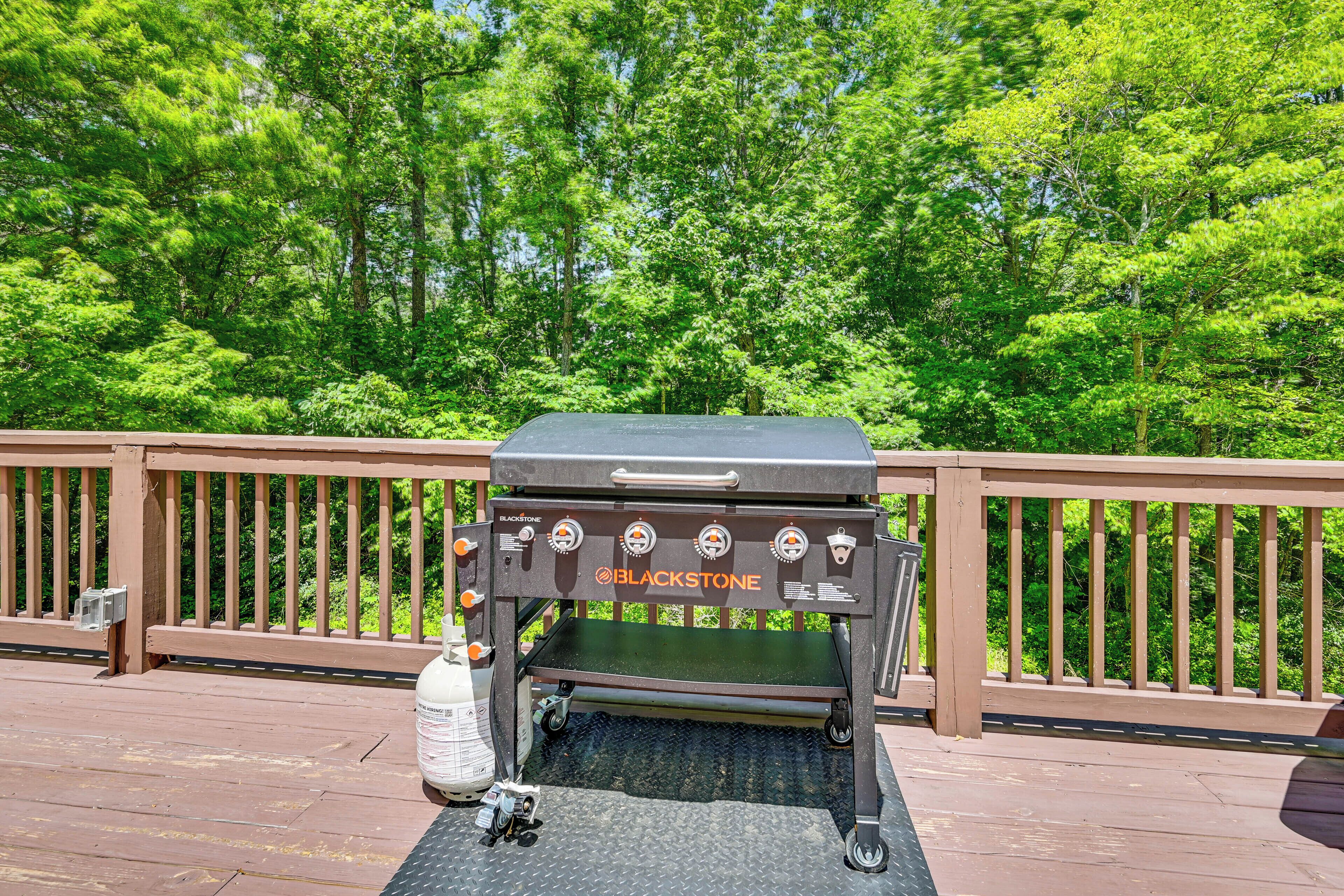Una parrilla Blackstone sobre una terraza de madera con rejilla metálica, rodeada de barandas de madera y árboles verdes y frondosos al fondo.