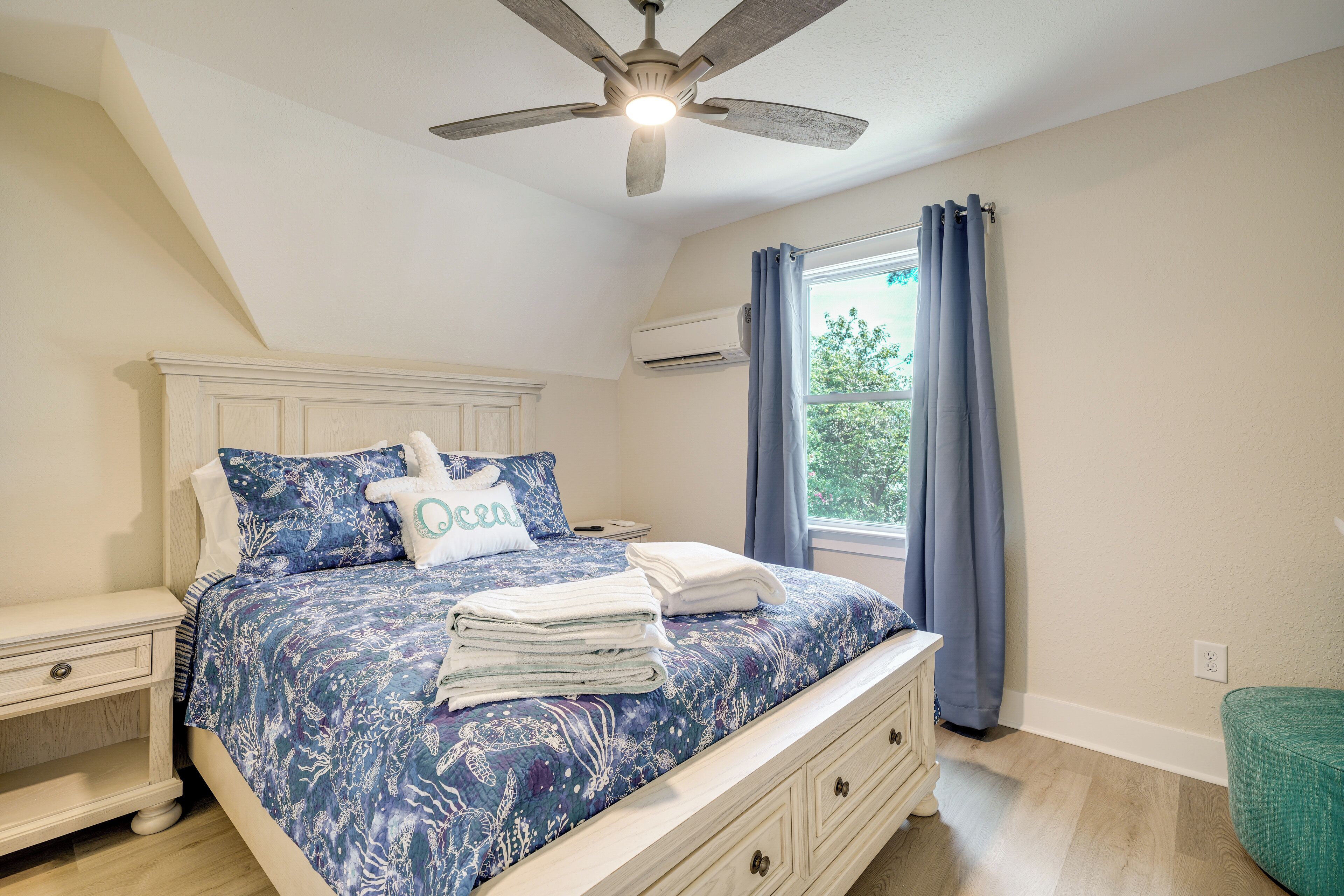 Un dormitorio con cama, mesitas de noche, ventilador de techo, ventana con cortinas y aire acondicionado.